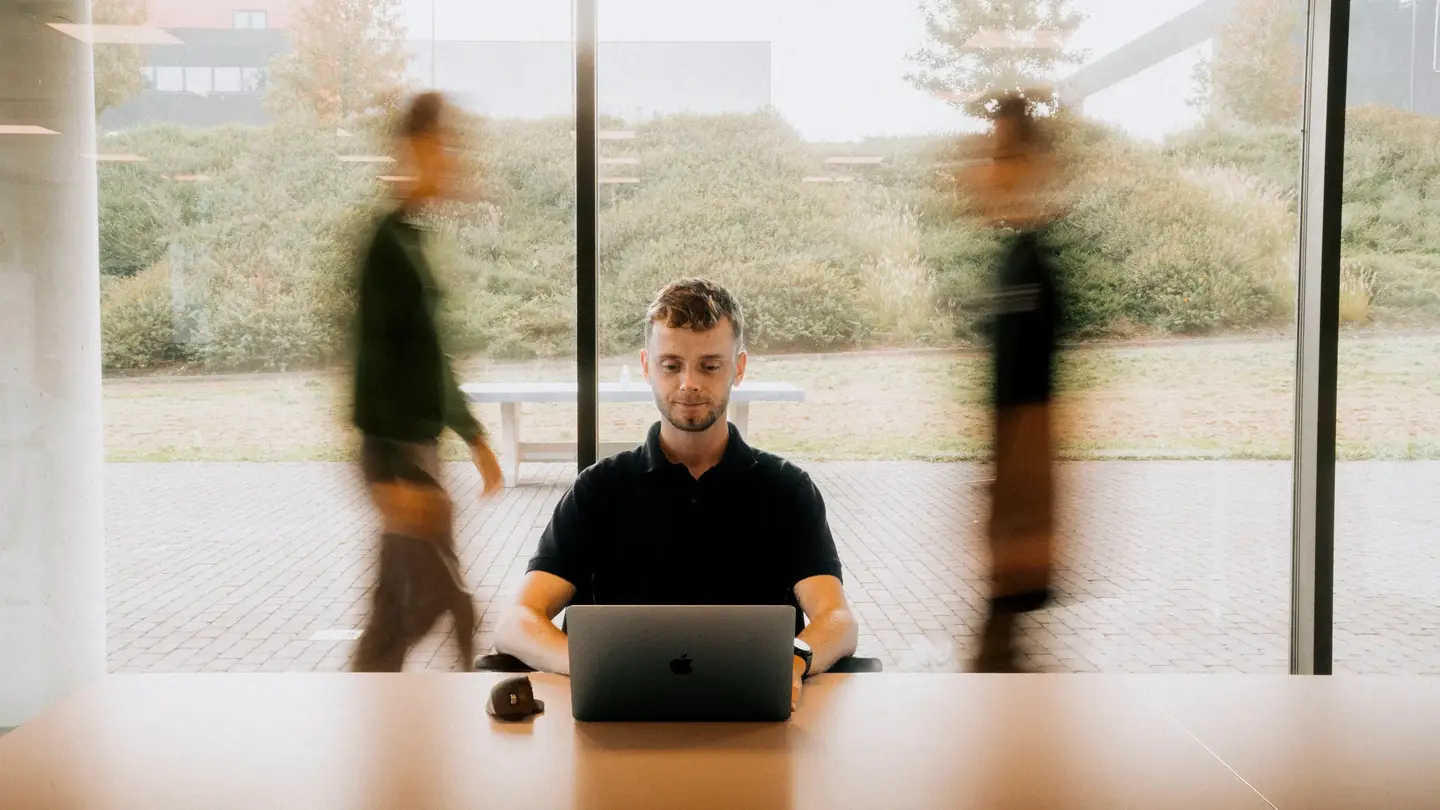 Un homme assis à une table dans une pièce éclairée travaillant sur un ordinateur portable. Deux silhouettes floues sont en mouvement derrière une vitre, créant un contraste avec la tranquillité de l'homme au premier plan.